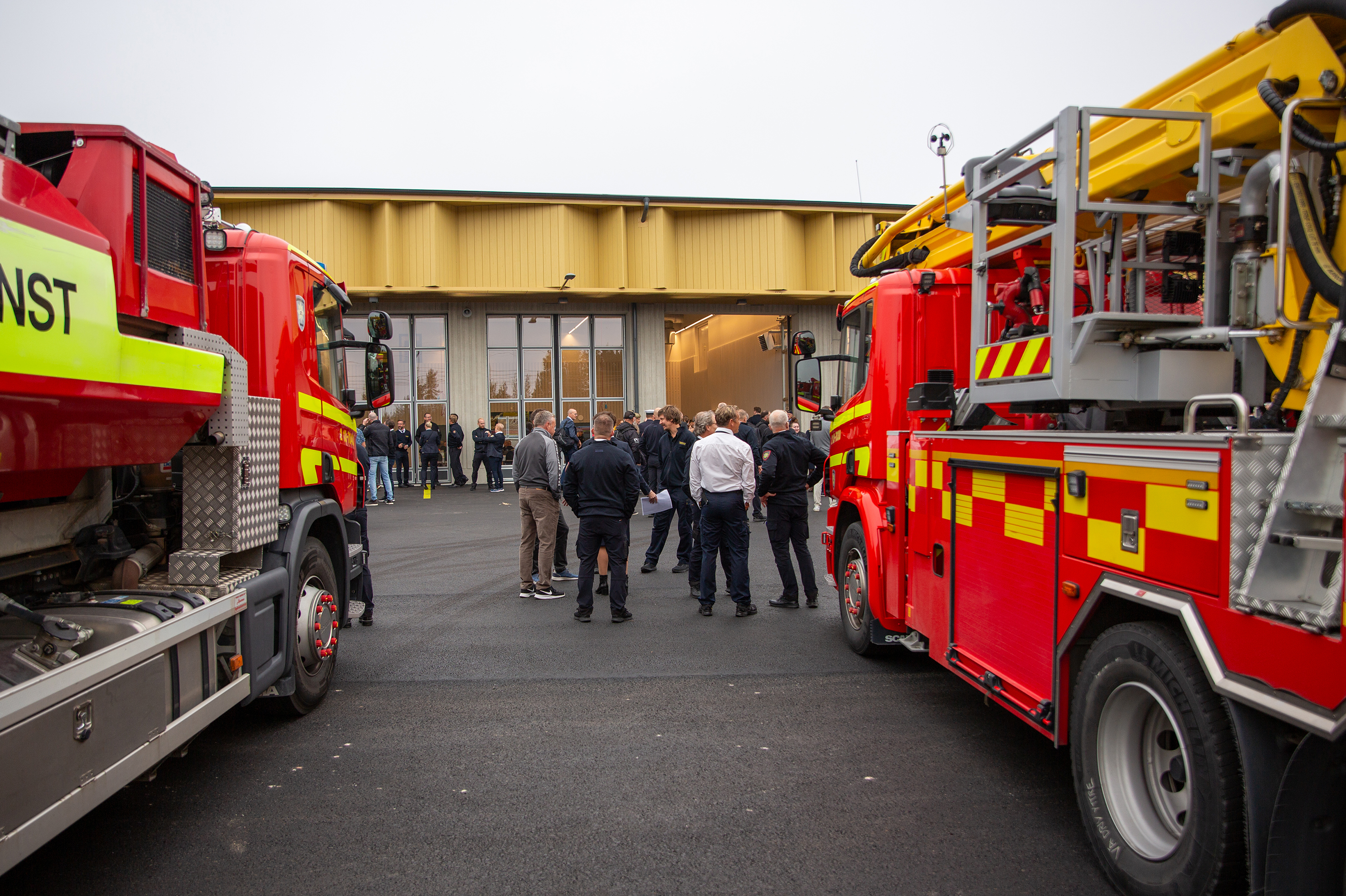 Två brandbilar syns i var sida av bilden. En grupp med människor står samlade framför den nya räddningsstationen i Timrå.